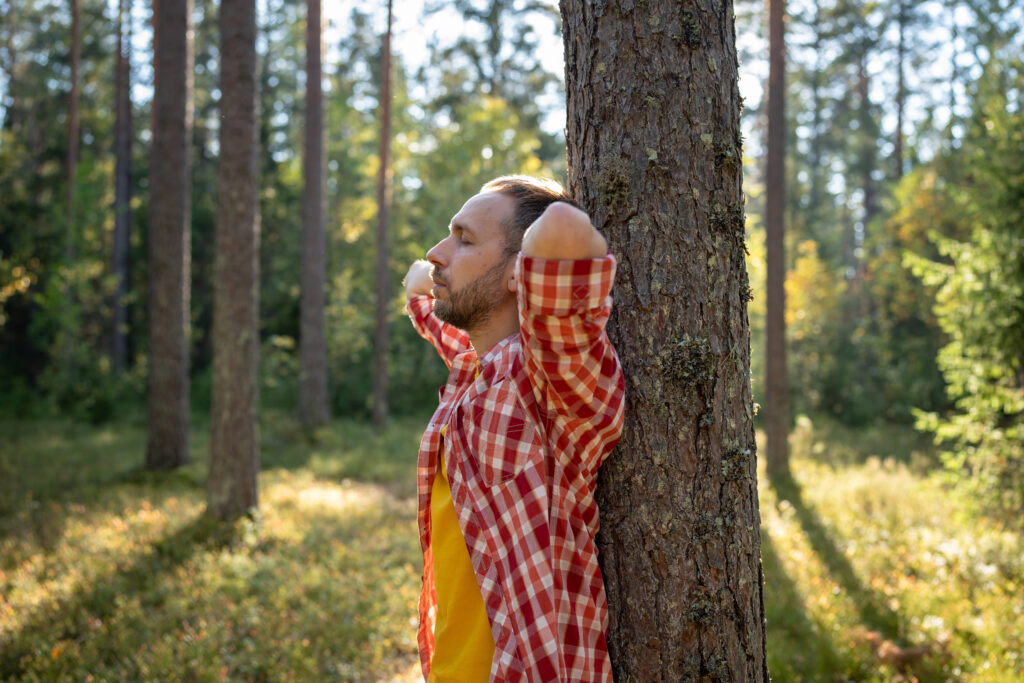 Man leans on pine tree standing in forest resting with closed eyes in hike. Relaxing in unity with nature feeling power harmony tranquility, spiritual practice experience. Guy meditating, zen state.